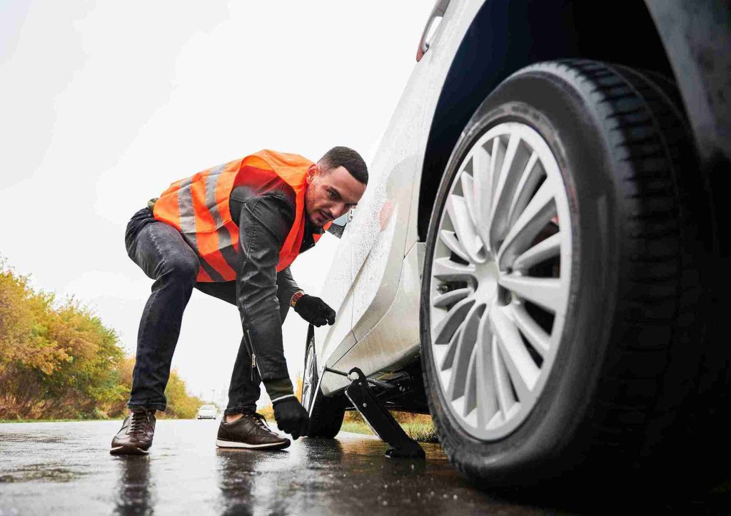 Roadside assistance technician changing a flat tire with a jack on a wet road in DeSoto TX